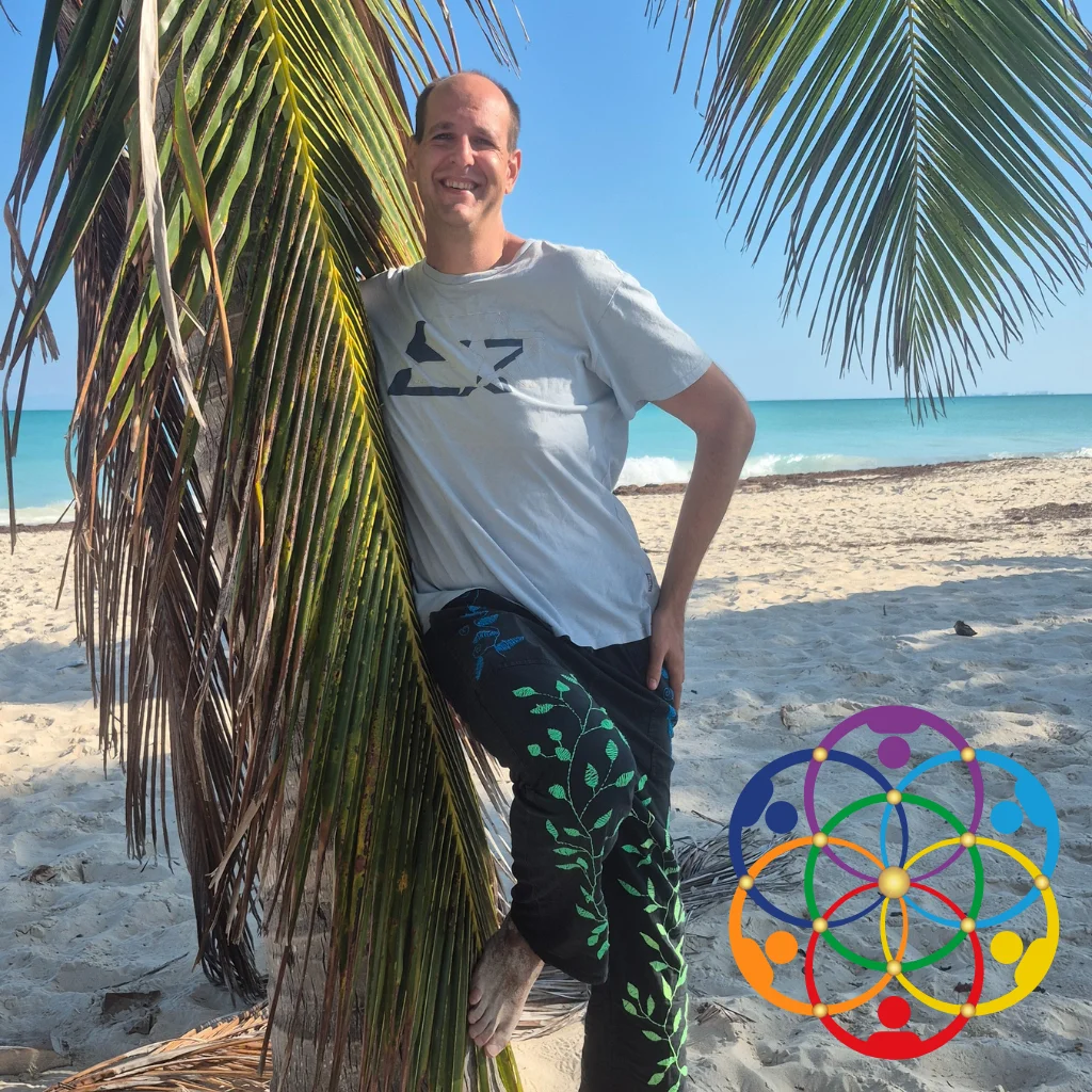Andreas Just - Über mich Bild Andreas Just steht barfuß an einem Strand neben einer Palme, trägt hellblaues T-Shirt und dunkle Hose mit grünem Pflanzenmuster, im Hintergrund Meer und blauer Himmel, unten rechts Logo mit Blume-des-Lebens-Symbol aus stilisierten Menschen in Chakra-Farben